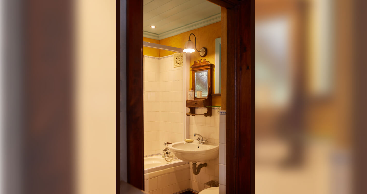 The quaint bathroom in Lover’s Nest cottage at TwoAngels. It has a  porcelain toilet, basin and bath. Above the basin is a rustic style wood framed mirror. The bathroom ceiling is a soft blue, and its walls are saffron.