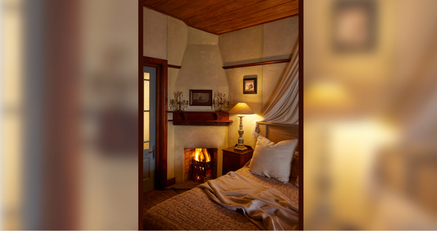 A fireplace burning next to the double bed in Lover’s Nest cottage at TwoAngels. The light in the room is warm and romantic. The bed is dressed in soft beige linen.