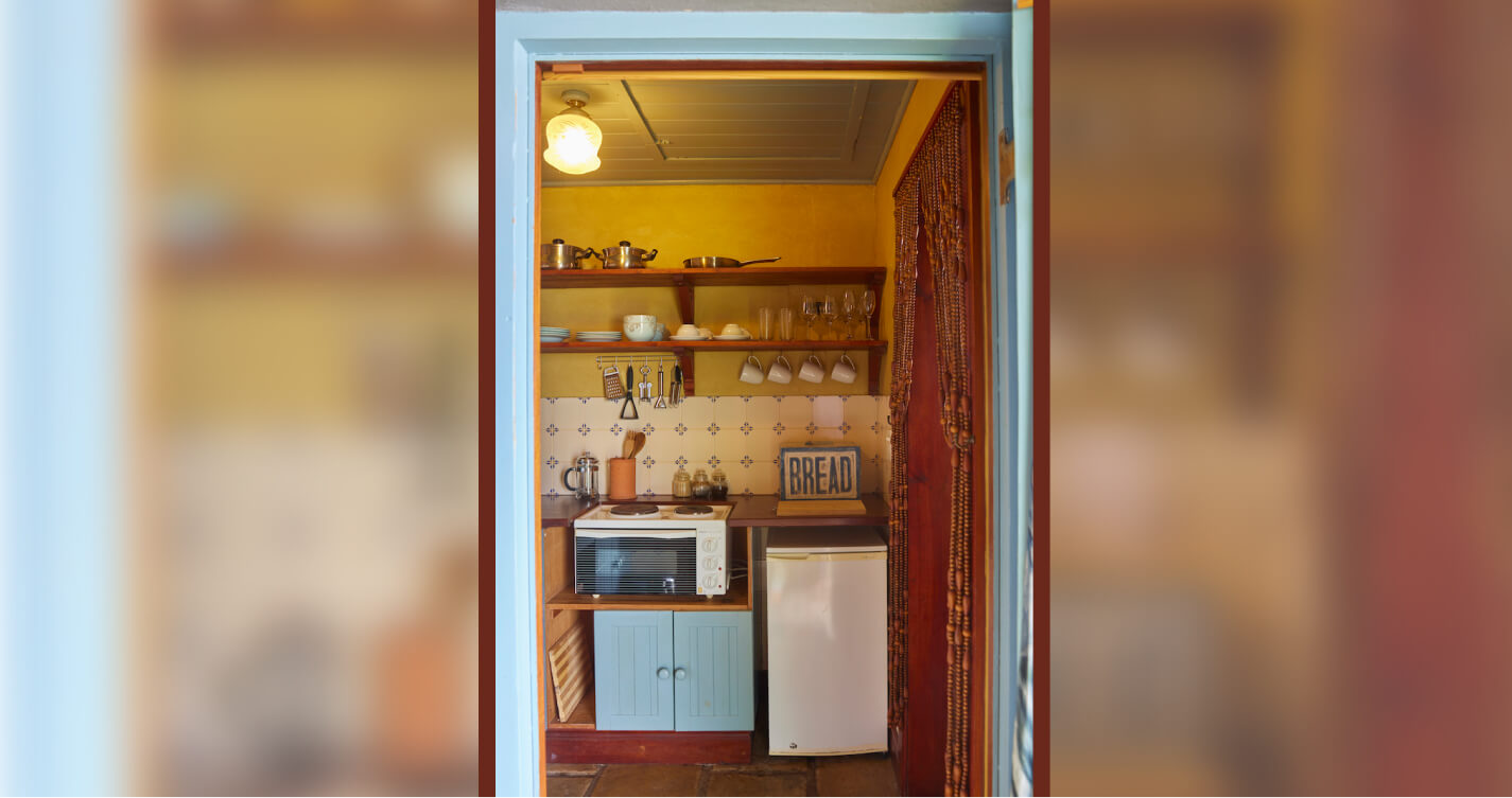 Looking  into the kitchenette of Lover’s Nest cottage at TwoAngels. The door frame is painted soft blue. The walls are saffron coloured. Built-in wooden shelves holding kitchenware sit above a wooden work surface. The work surface wraps around a two hob stove and a little bar fridge.