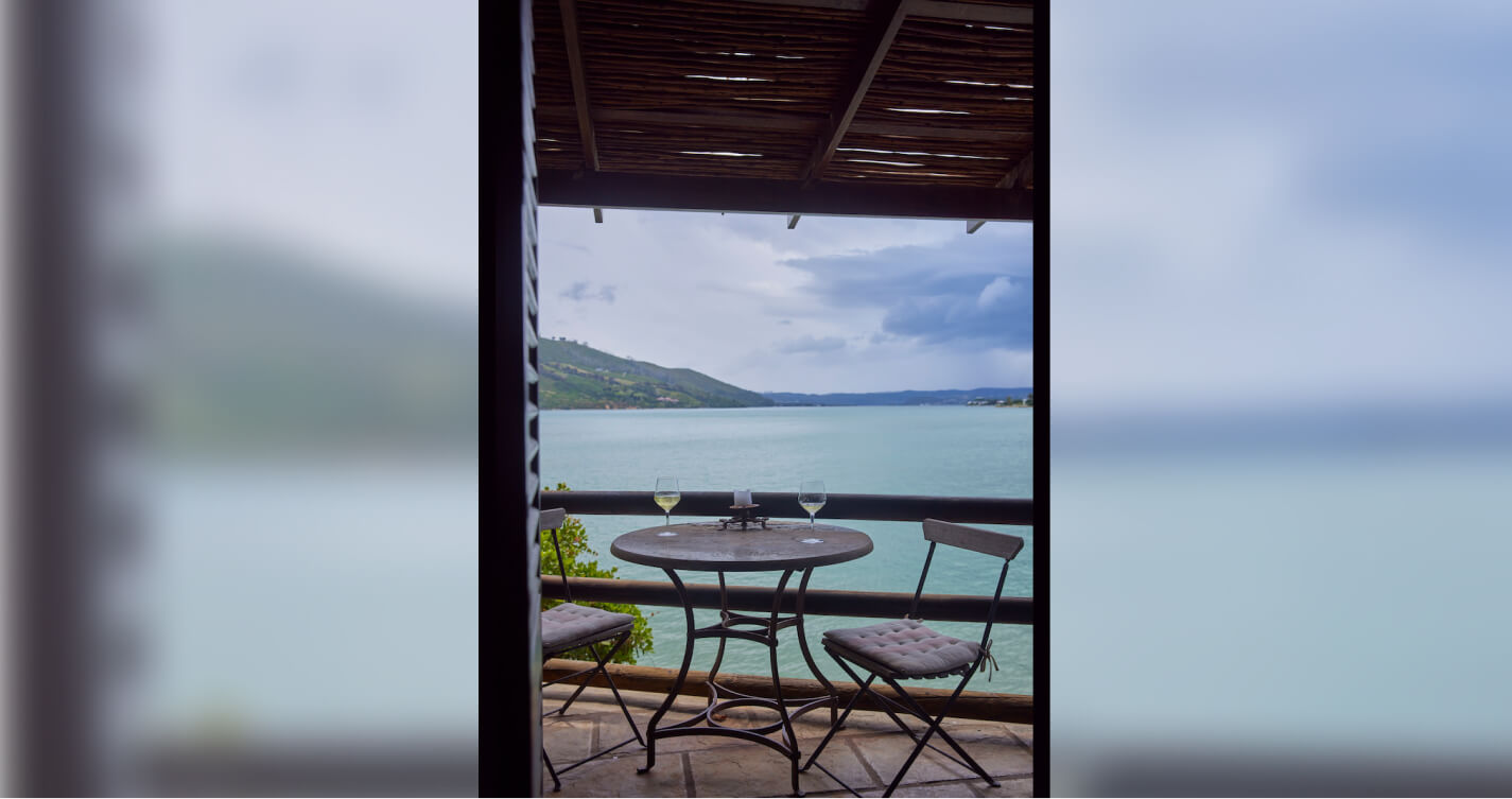 The seaside balcony in Lover’s Nest cottage at TwoAngels. On the balcony is a table for two. Two glasses of white wine stand on the table. The light is soft and calm. Beyond the table is the sea, a mountain range and a dramatic cloud decorated sky.