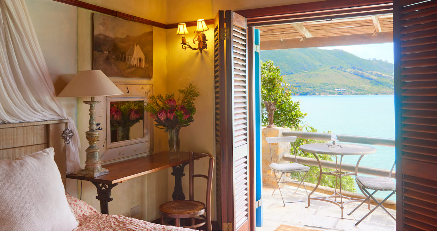 A romantic sea view from the bed of Lover’s Nest cottage at TwoAngels in Knysna. The bed is in the foreground. It looks out through open french doors, over a quaint private balcony with a table for two, and onto a body of blue water.