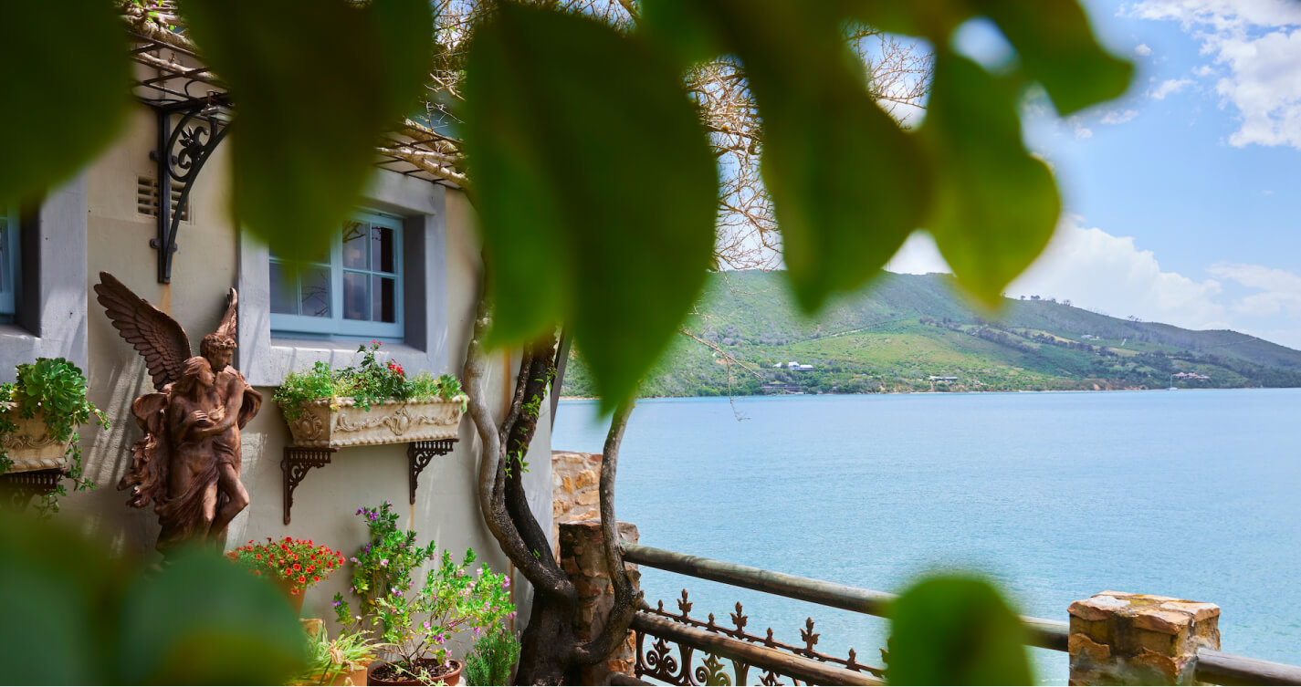 A warm sunny day at TwoAngels in Knysna. Lover’s Nest cottage stands next to the sea. The cottage has blue window frames and is surrounded by flowers. A sculpture of two angels stands in the flower bed. The sea water is blue and beyond it the southern head mountain is visible.