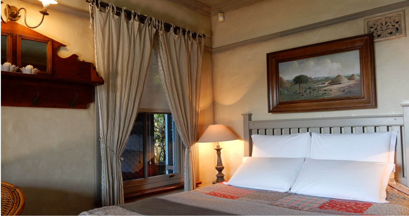 The Oyster Shell cottage’s double bed stands next to an open window. The window allows a soft, relaxing light to enter the room through thin  linen curtains. The rooms walls are a calming butternut colour. A wooden framed artwork decorates the wall at the head of the bed.