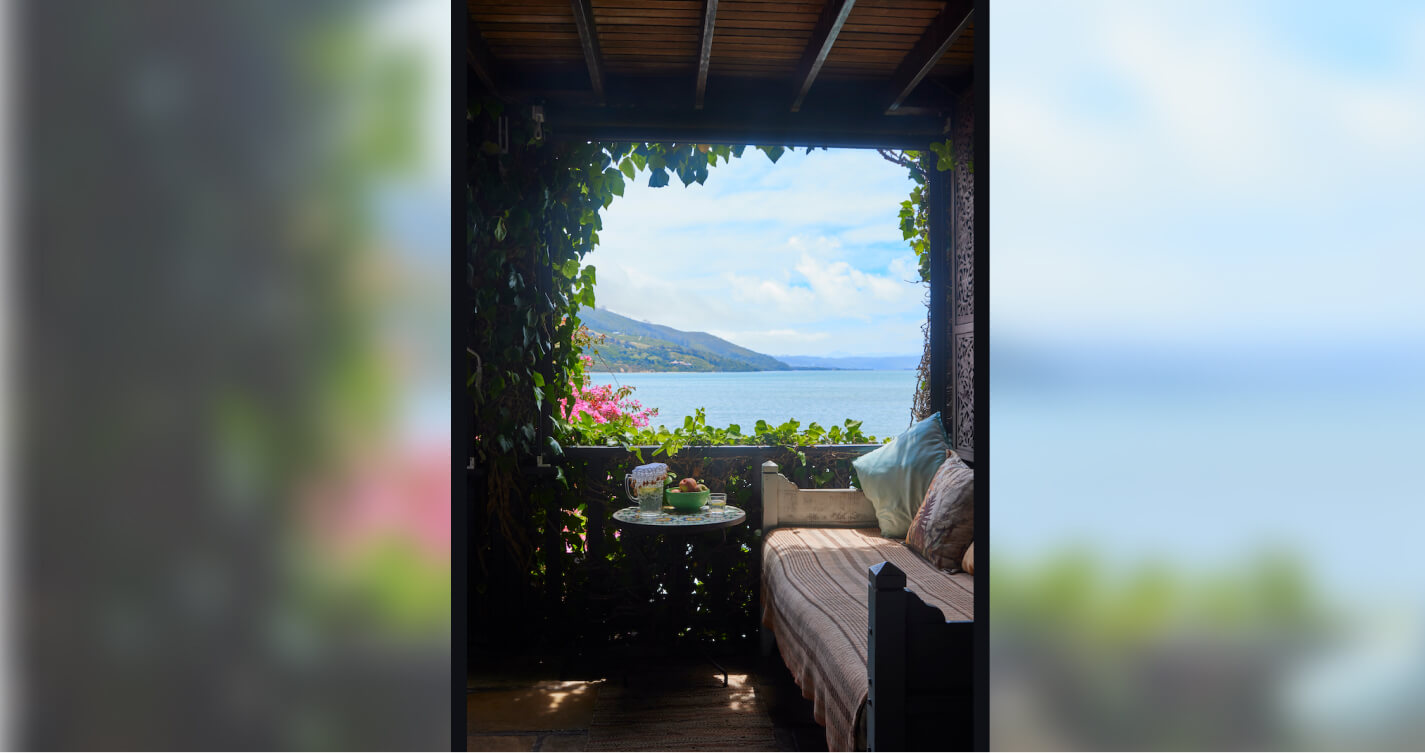 A relaxing looking daybed with a sea view. The daybed is against one wall of an enclosed wooden balcony. The view is framed by the green leaves of a bougainvillea that creep around the balcony structure. The water is blue and sun lit.