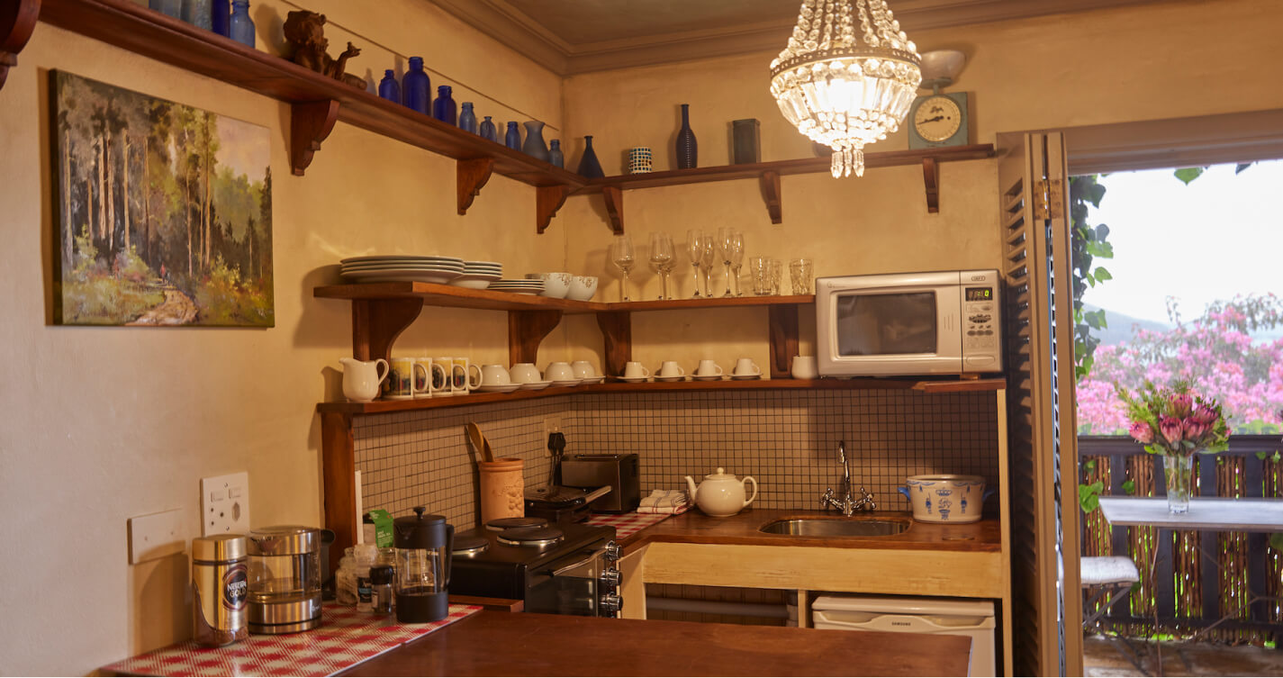 The Oyster Shell cottage’s open plan kitchenette. The kitchenette has built-in wooden shelves containing kitchenware. Below the shelves is a  U shaped wooden work surface containing a stove, sink and bar fridge.
