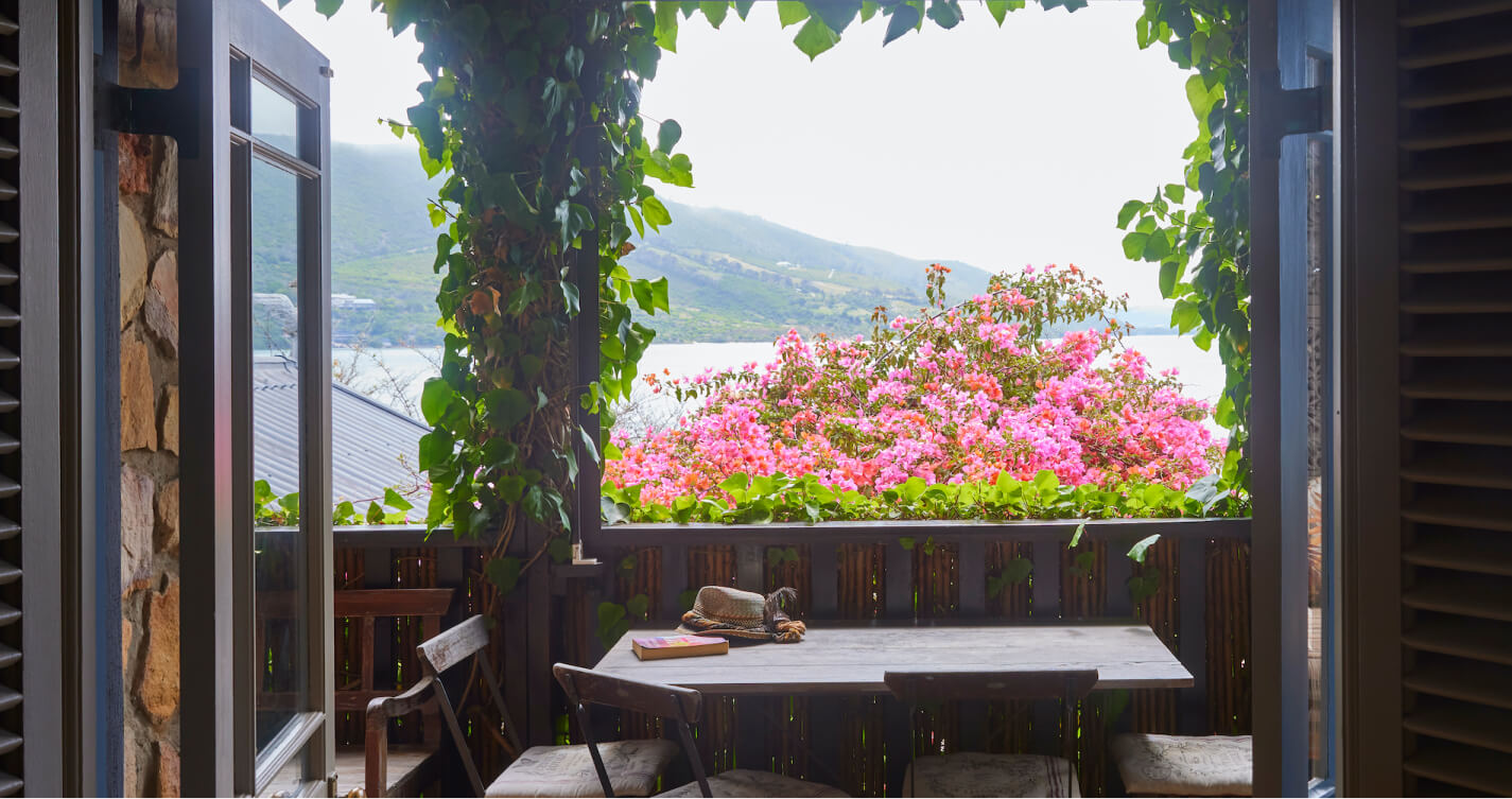 Looking out through the open french doors of the TwoAngels Oyster Shell cottage. There is a wooden dining table and chairs.  The view  beyond the table is of a Bougainvillea, surrounded by sea, mountains and sky.