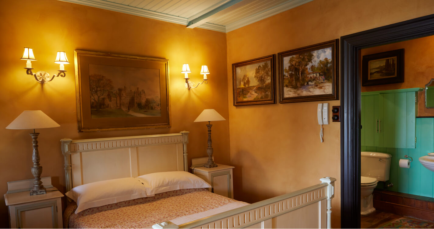 The Owls Nest bedroom. Saffron walls decorated in landscape paintings surround a double bed. Through a doorway the en suite bathroom is visible. The bathroom is painted in burnt orange and shades of green.