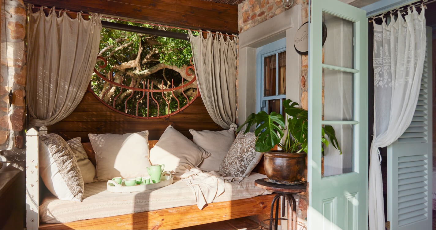 The Owls Nest bedroom daybed. It is tucked into a corner of the bedroom's private balcony. It is covered in pillows, creating the perfect place to relax. Gentle sunlight streams onto the bed and healthy pot plants grow at its side.