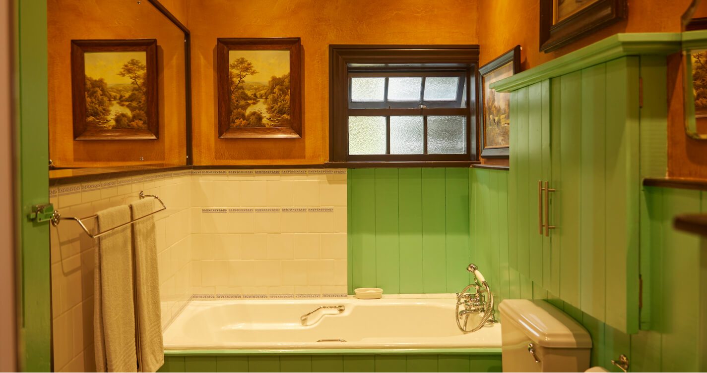 The beautiful en-suite bathroom of Owls Nest bedroom. A window fills the bathroom with natural light. The walls are painted in colours of green and burnt orange.  Landscape paintings hang on the walls above the bath. Another copy of one of the paintings is reflected in a large mirror.