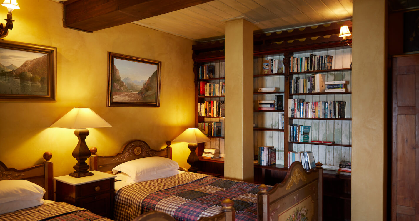 The Robbins Nest bedroom in the TwoAngels Family Suite. It has two single beds. The light is calming. The beds are separated by a bedside table. There is a landscape painting hung above each bed. One of the walls is a well stocked book shelf.