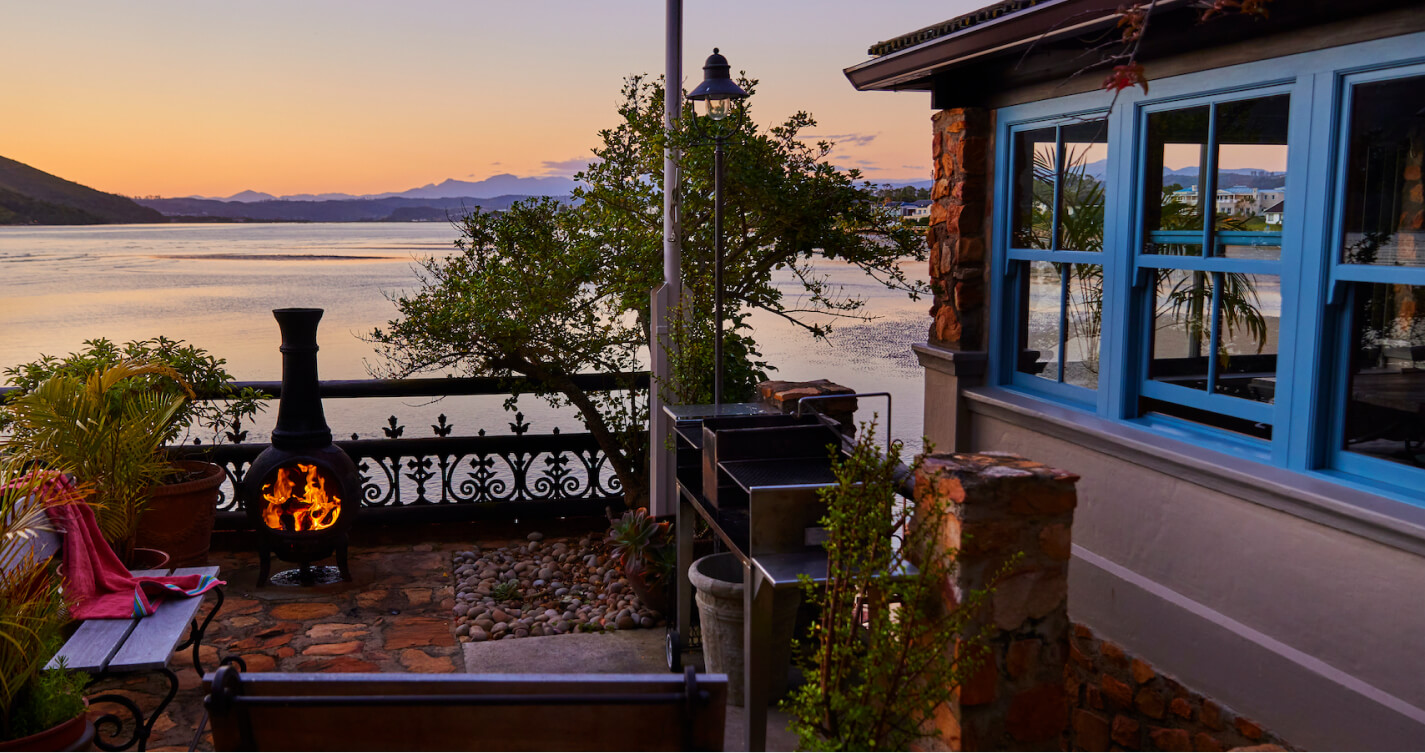 The Family Suite braai area. It offers a magical sea view. Two wooden benches allow seating around the braai. The surrounding garden flourishes with healthy plants. An eye catching fire burns as the sky is filled with colours from the setting sun.