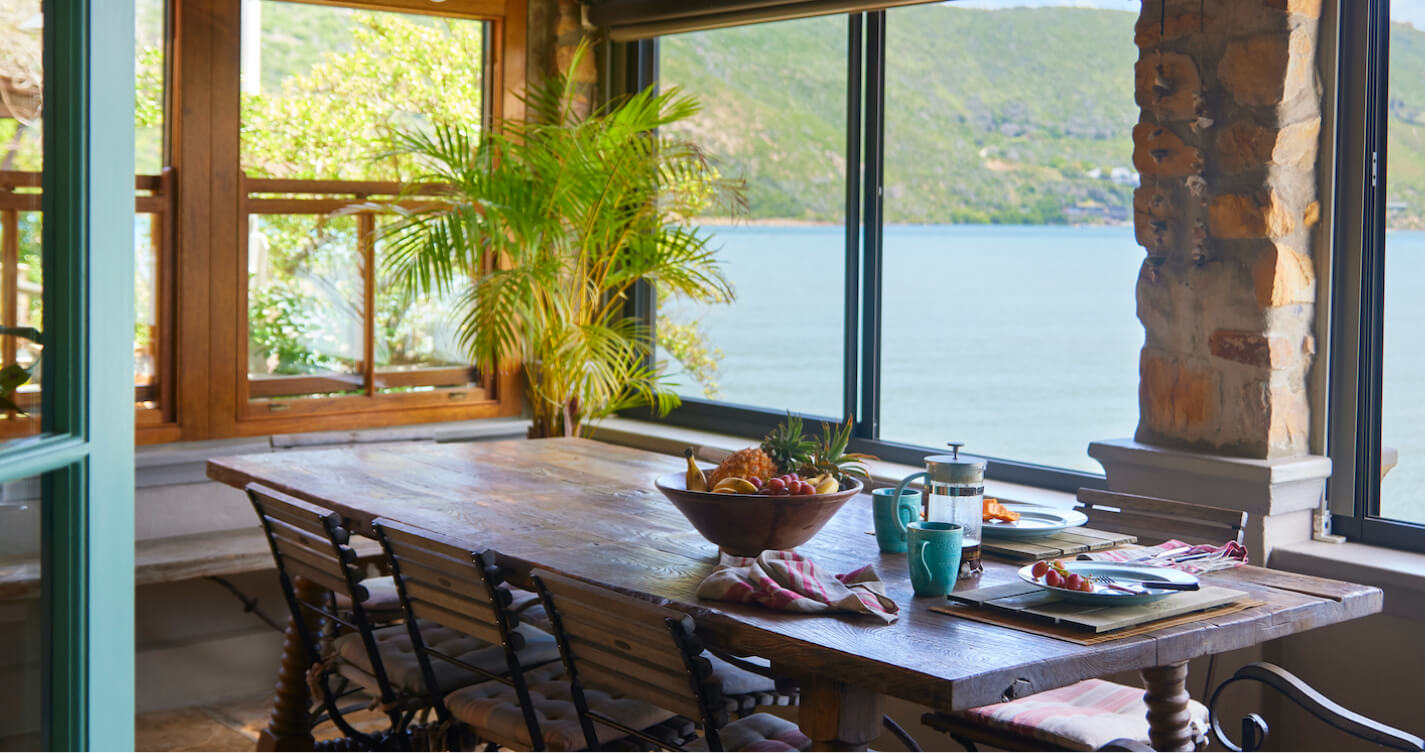 The TwoAngels Family Suite dining room with its sea view. Natural light fills the room through surrounding windows. A big wooden dining table looks out onto a sea of blue water. There is a bowl of colorful fruit on the table.