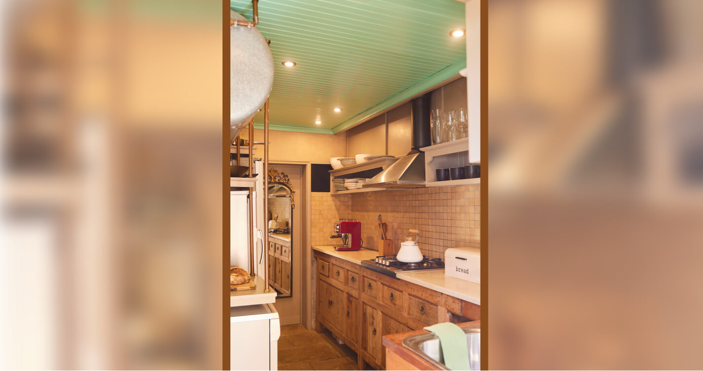 The Family Suite Kitchen. The roof is green. A marble counter runs along one wall for the length of the room. A gas stove is built into a section of the counter. The kitchen cabinets below the counter are wooden. Visible in the kitchen is a fresh loaf of bread, a coffee machine, a kettle and a sink.