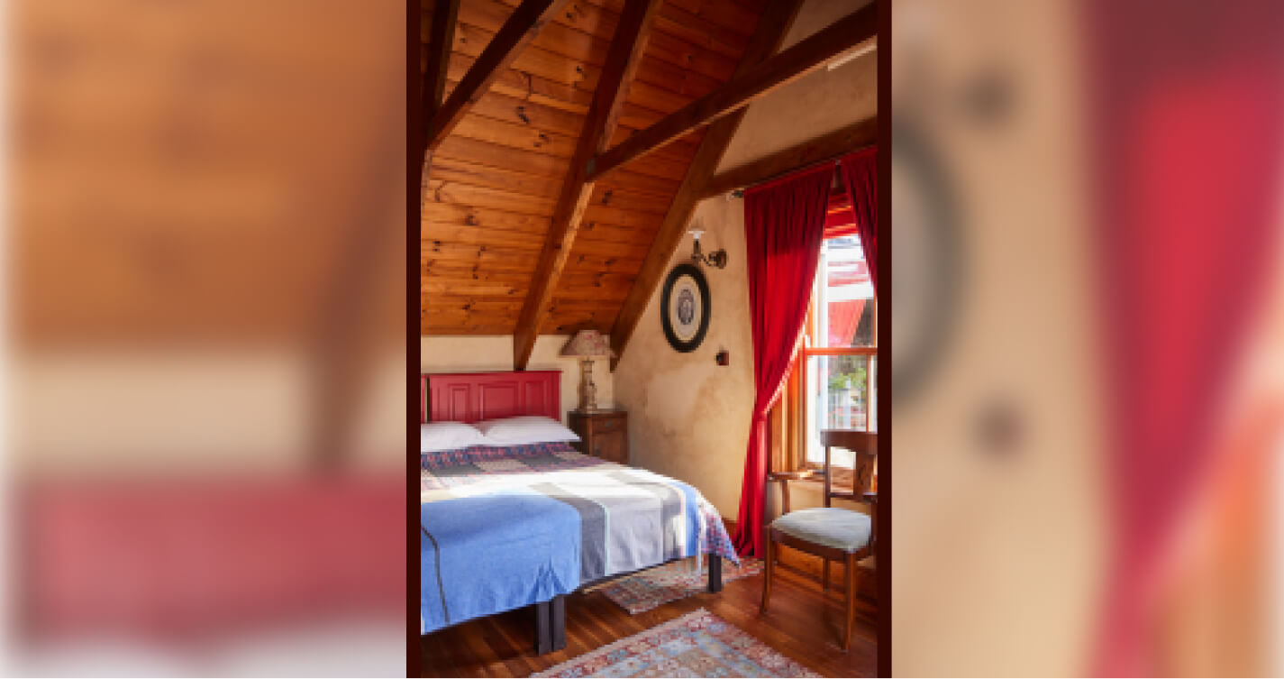 The Madonna bedroom, bright with natural light. A wooden chair with a green cushioned seat stands near the window. The double bed is dressed in blue, gray and crimson bedding. The floors are wooden and decorated by patterned rugs.