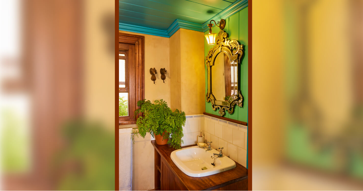 Madonna bedroom’s en-suite bathroom. It is quaint, painted green and blue and filled with natural light. A flourishing fern stands near the open window. The basin is set into a wooden cabinet. A mirror above the basin reflects the frame of the window.