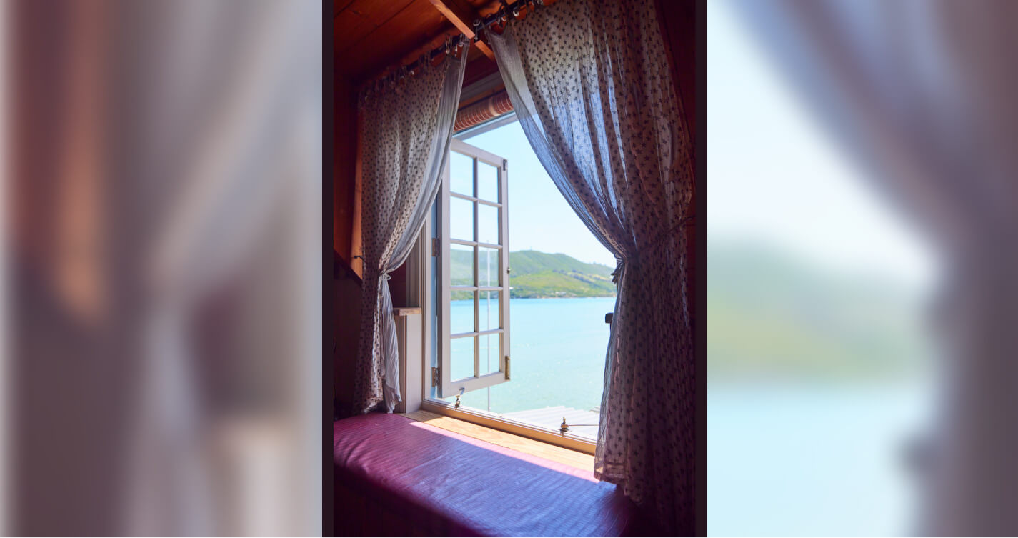 A romantic sea view from the Madonna bedroom in the TwoAngels Luxury Suite. Two light linen curtains hang on either side of an open window. Outside the sky is bright and the water is blue. Inside the light is soft and calming. A wooden bench with a burgundy seat is directly in front of the window.