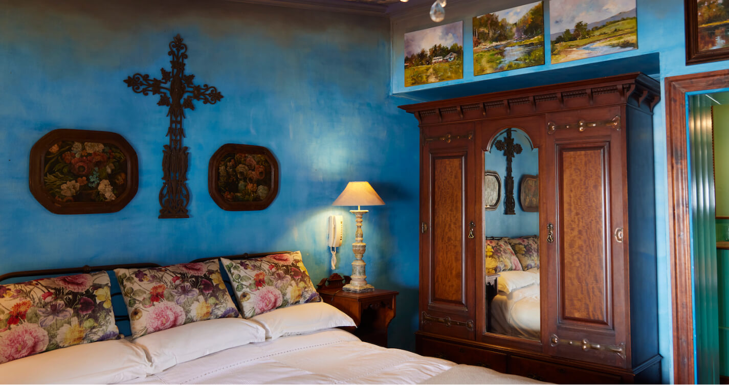 The Luxury Suite Sunset bedroom. Blue walls surround a double bed. At the head of the bed are three decorative rose pillows. A large wrought iron cross hangs on the wall behind the bed.