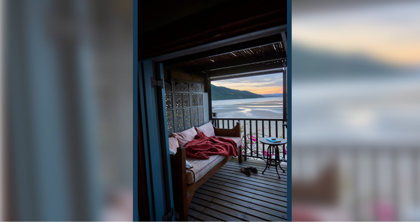The private deck of the Sunset bedroom. On the deck is a daybed with a sea view. It’s late afternoon and the sun is setting. The sky is orange, yellow and blue. The water below reflects the colours in the sky.