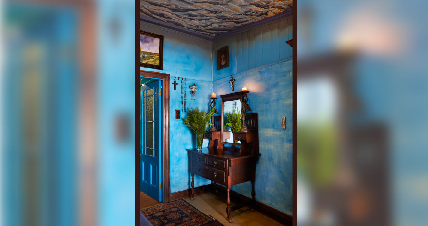 A dark wooden dresser with a mirror stands in the corner of  the blue walled Sunset bedroom. On the dresser is a vase of fynbos and two carved angel candelabras.