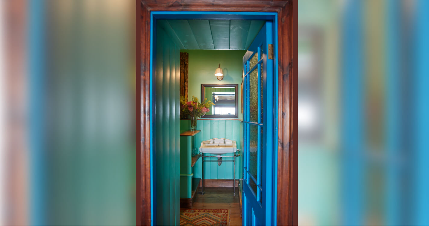 Looking through a doorway at the mirror and basin in a turquoise and blue bathroom. It is the en-suite bathroom of the Sunset bedroom.
