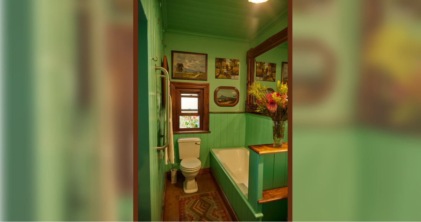 The en-suite bathroom of the Sunset bedroom has green walls decorated with landscape paintings. There is an open window behind the toilet, and an enormous mirror on the wall above the bath. A decorative rug runs the length of the wooden floor.