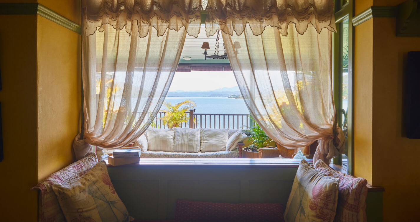 The tranquil feeling Luxury Suite living room with its sea view. In the background is a body of blue water. It's visible through an open window that stretches the length of the entire room. Natural light pours into the room and onto couches with lots of pillows. Thriving pot plants grow next to the couches. A thin linen curtain softens the light.