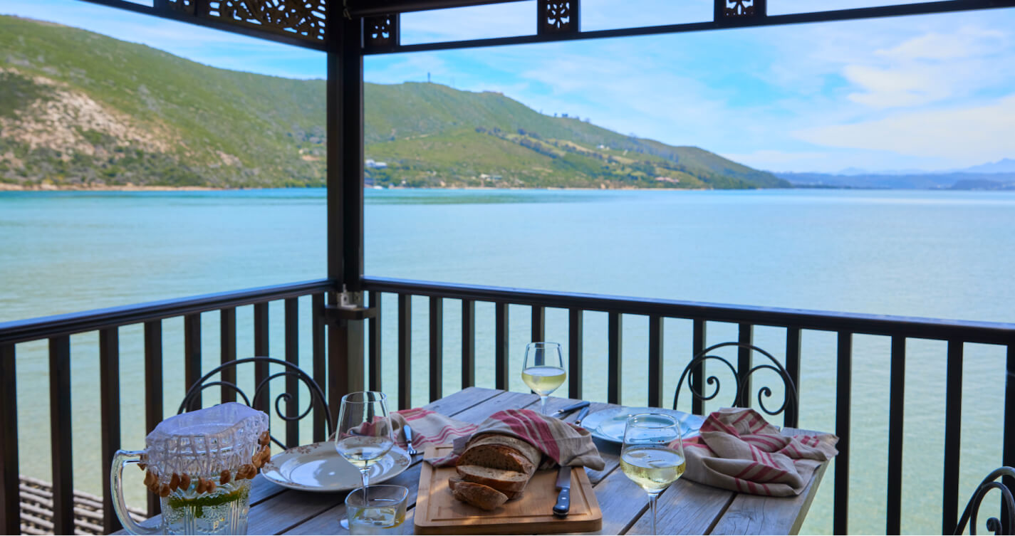 The TwoAngels Luxury suite dining table on its balcony with its sea view. In the foreground is the table. It contains a wooden chopping board with a freshly sliced loaf of bread and three glasses, each with a varying amount of white wine. In the background is a body of blue water surrounded by mountains and sunny sky.