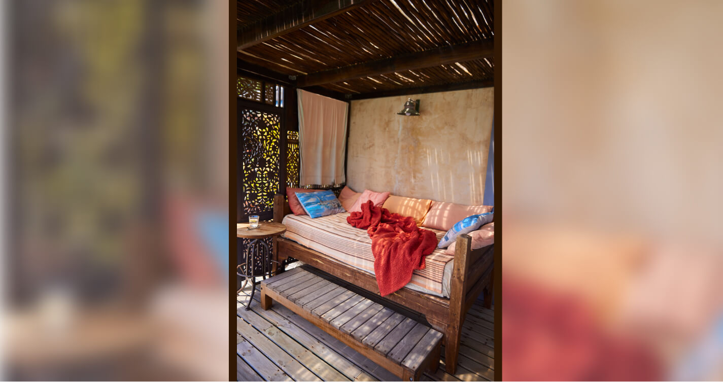 The daybed on the Luxury Suite balcony. It is covered by sapling weaved roof that allows through dappled light. The light falls onto the burnt oranges and blues of the bed and all its pillows. It looks like a great place to relax with a book or take a nap.