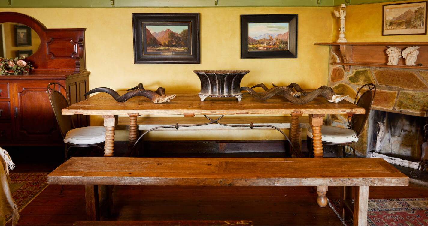 The Luxury Suite dining room with its large wooden table next to its stone fireplace. Two matching wooden benches run the length of the table. At each head is a wrought iron chair. Landscape paintings hang on the surrounding saffron coloured walls. The floor is wooden with areas covered by decorative rugs.