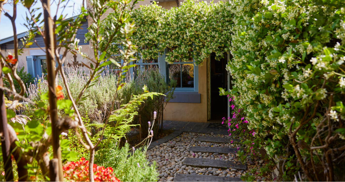 The TwoAngels garden blossoms with jasmine flowers, lavender bushes, ferns and begonias. A bed of pebbles with a walkway made from wooden planks runs through the middle of the garden. The walkway ends at the entrance to the Luxury Suite.
