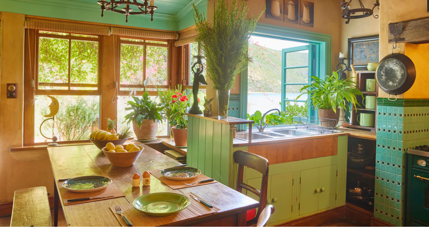 The large TwoAngels Luxury suite kitchen. It is surrounded by windows that fill it with a warm, natural light. A wooden dining table surrounded by benches and chairs is set to the one side. Pot plants and bowls of fresh fruit pop with colour. Surfaces painted green, blue and saffron compliment the sea and mountain beyond the windows.