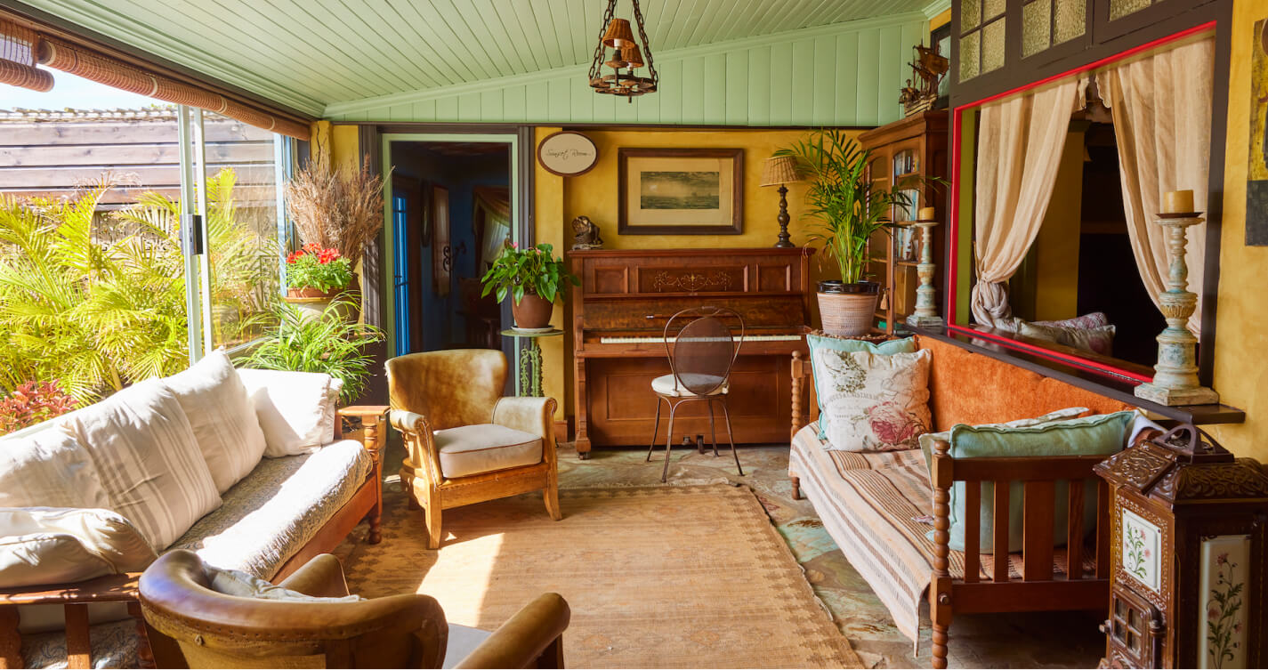 Natural light floods into the Luxury Suite living room. It enters through a large sliding window. Beautiful pot plants bring life to the room. Comfortable looking couches packed with pillows offer space to relax. A piano stands against one wall of the room.