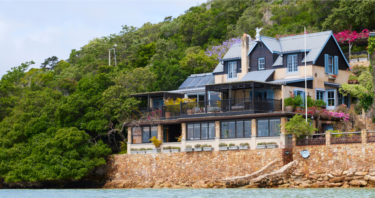 TwoAngels guest house on the water's edge. The house is set into a backdrop of trees. It has a tanned wooden facade with a sky blue roof and windows. At the base of the house is a stone wall with a jetty that leads into a body of blue water.