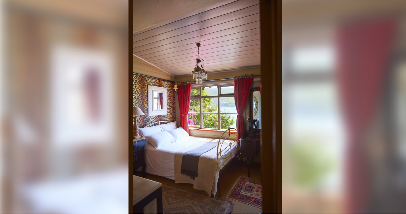 The Angel’s Lair master bedroom. It has a double bed and a sea view. The bed runs along a large, sea facing window. At the head of the bed is a leopard print wall with a small mirror hung at its center. A crystal chandelier hangs above the bed. Decorative rugs bring color to the carpeted floor.