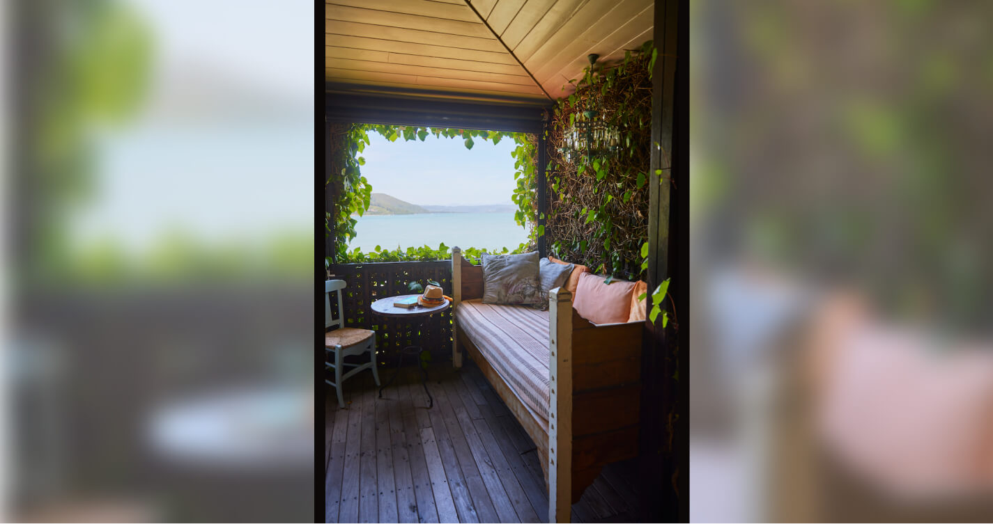 The romantic sea view from the Angel’s Lair balcony at TwoAngels in Knysna. In the background is a sunny sky, mountains and blue water. In the foreground is a comfortable looking daybed with lots of pillows. The green leaves of a Bougainvillea surround the balcony and frame the view.