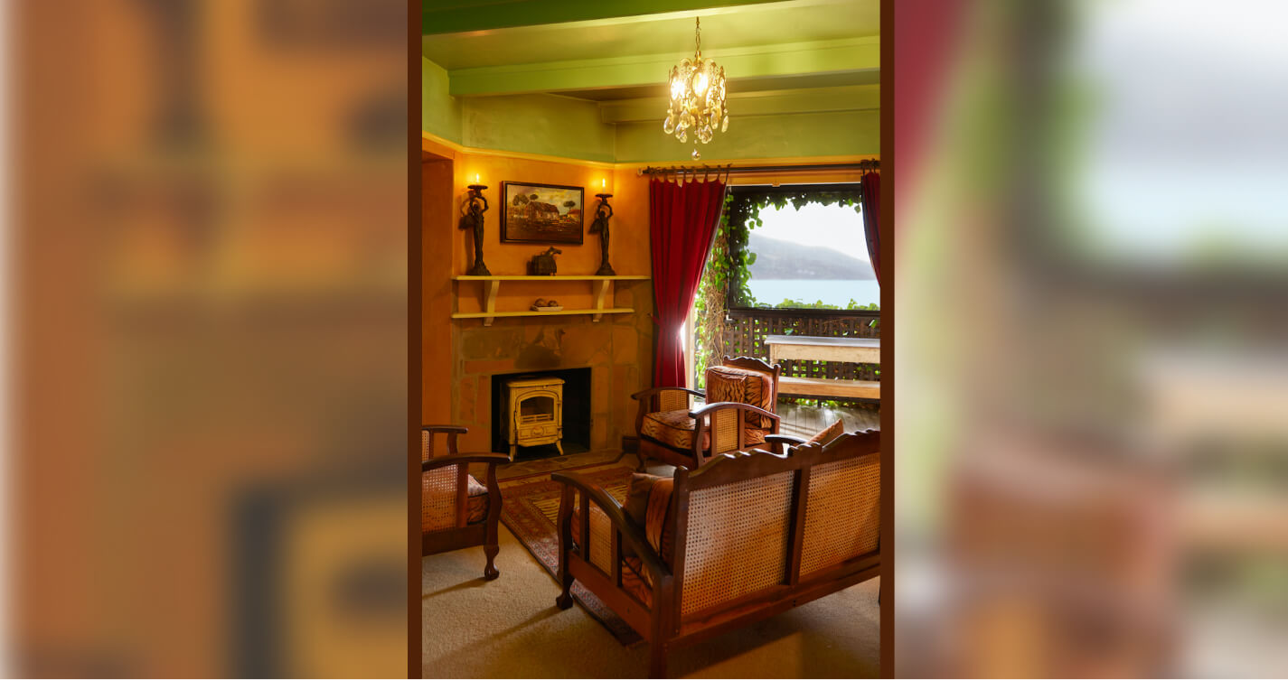 The Angel’s Lair has a cozy, open plan living room with a sea view. Three couches face into the center of the room. They look onto a wall with a built-in fireplace. There is a carpet beneath them. Double french doors lead out onto a sun lit balcony. There is a sea of blue water beyond the balcony.