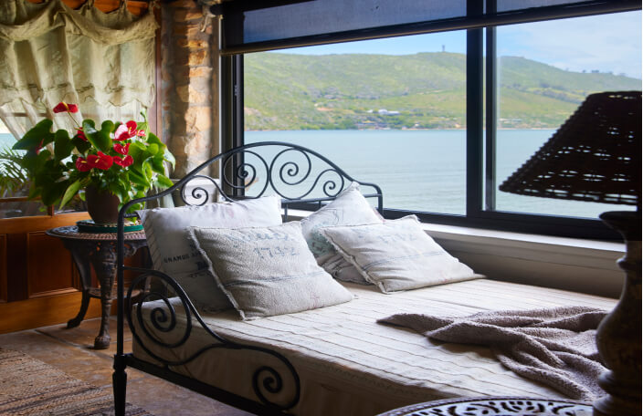 A comfortable looking daybed on Family Suite's covered balcony. The bed stands next to large windows that offer a sea view. It looks like the perfect place to relax.