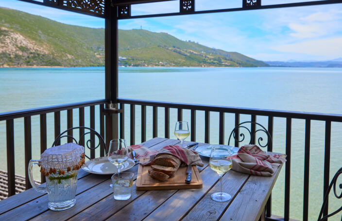 The TwoAngels Luxury suite dining table on its balcony with its sea view. In the foreground is the table. It contains a wooden chopping board with a freshly sliced loaf of bread and three glasses, each with a varying amount of white wine. In the background is a body of blue water surrounded by mountains and sunny sky.