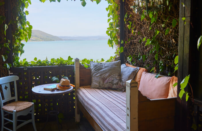 The romantic sea view from the Angel’s Lair  balcony at TwoAngels in Knysna. In the background is a sunny sky, mountains and blue water. In the foreground is a comfortable looking daybed with lots of pillows. The green leaves of a Bougainvillea surround the balcony and frame the view.