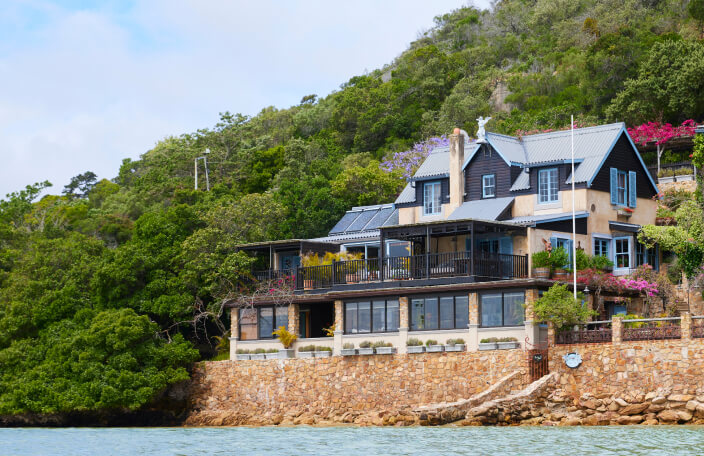 TwoAngels guest house on the water's edge. The house is set into a backdrop of trees. It has a tanned wooden facade with a sky blue roof and windows. At the base of the house is a stone wall with a jetty that leads into a body of blue water.